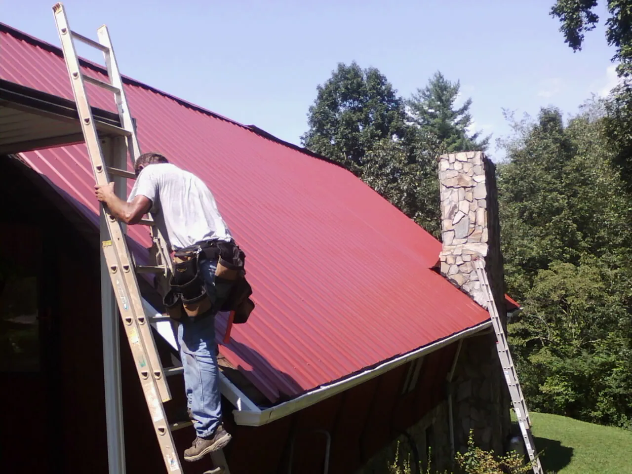 Roofer on a ladder beside a red metal roof and stone chimney — Soffit & Fascia Repair by Roofing Brothers in Schwenksville